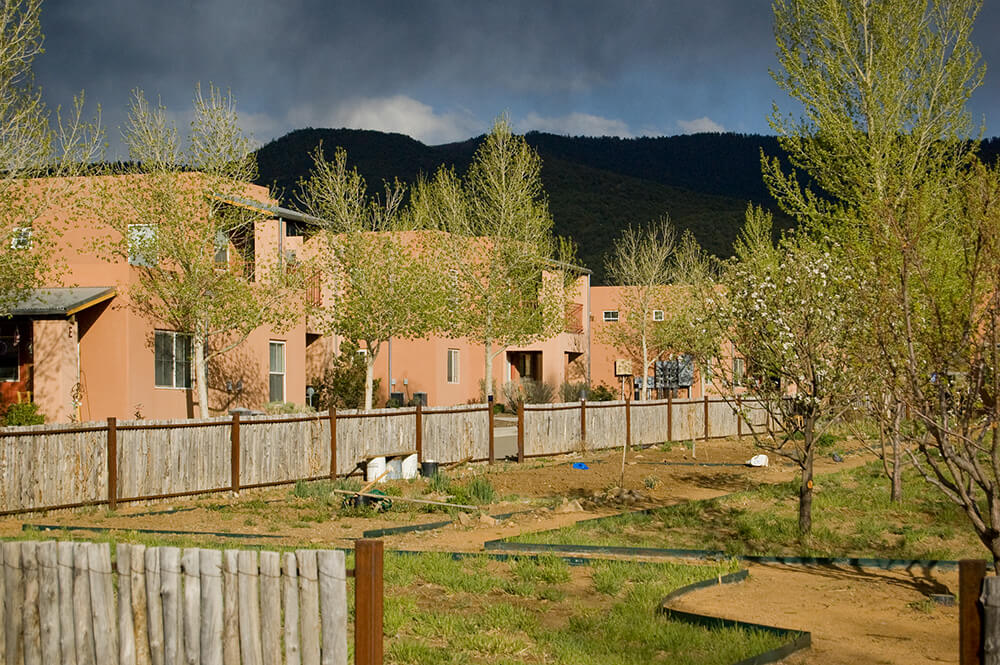 Golden evening light on Loma Parda buildings with the dark forested mountainside behind, Taos NM